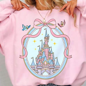 May include: A light pink sweatshirt featuring a cartoon castle design within a pink ribbon and pearl frame. Two cartoon birds are on either side of the ribbon. The castle is pink and blue with yellow stars.