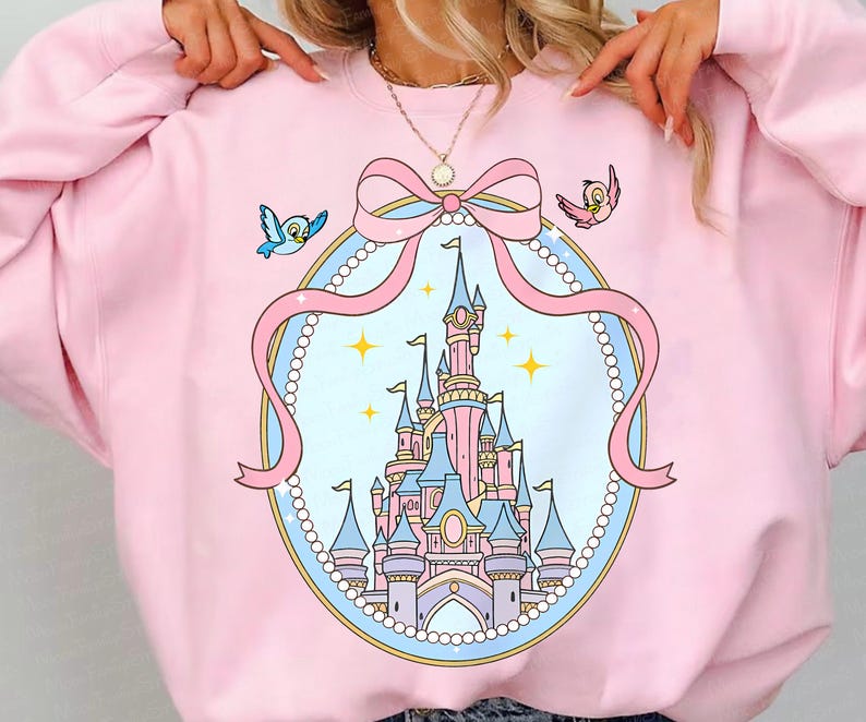 May include: A light pink sweatshirt featuring a cartoon castle design within a pink ribbon and pearl frame. Two cartoon birds are on either side of the ribbon. The castle is pink and blue with yellow stars.