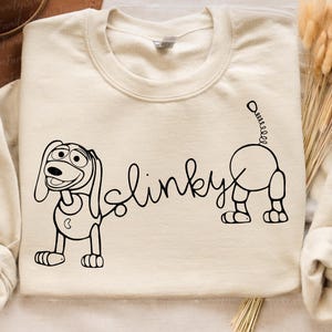 Peut inclure: Un sweat-shirt cr&egrave;me avec un dessin au trait noir de Slinky Dog de la franchise Toy Story. Le dessin comprend les mots "Slinky" et une repr&eacute;sentation du corps et de la t&ecirc;te de Slinky Dog.