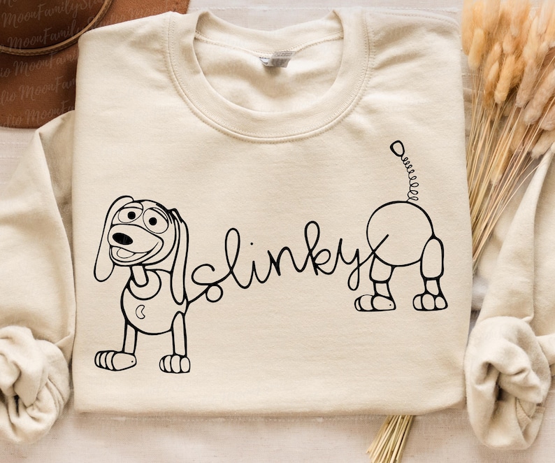 Peut inclure: Un sweat-shirt cr&egrave;me avec un dessin au trait noir de Slinky Dog de la franchise Toy Story. Le dessin comprend les mots "Slinky" et une repr&eacute;sentation du corps et de la t&ecirc;te de Slinky Dog.