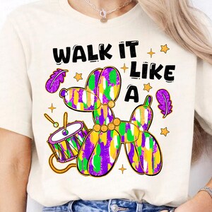 May include: Cream-colored t-shirt with a colorful graphic. The graphic features a balloon dog in purple, green, and yellow, a drum, and the text "WALK IT LIKE A". The design includes stars and purple feathers.