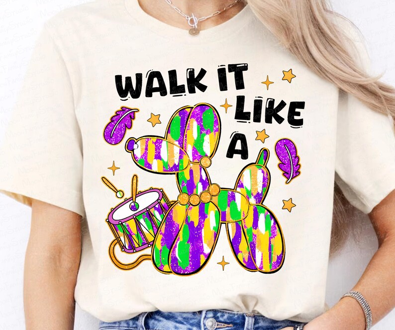 May include: Cream-colored t-shirt with a colorful graphic. The graphic features a balloon dog in purple, green, and yellow, a drum, and the text "WALK IT LIKE A". The design includes stars and purple feathers.