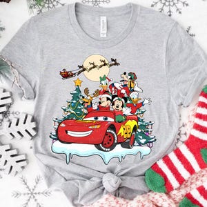 Pu&ograve; includere: T-shirt grigio chiaro con una grafica festiva di personaggi Disney in un'auto da corsa rossa, con un albero di Natale e la slitta di Babbo Natale sullo sfondo. Il design include Topolino e Minnie, Paperino e Pippo.
