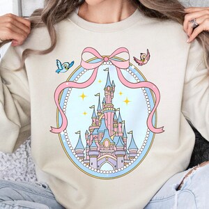 May include: Beige sweatshirt featuring a pastel castle design within a pink ribbon-framed oval. Two small birds flank the ribbon. The castle is pink, blue, and purple, with gold accents. The design is whimsical and cartoon-like.