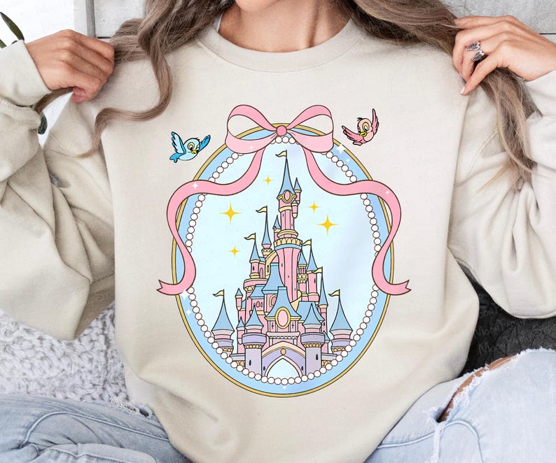 May include: Beige sweatshirt featuring a pastel castle design within a pink ribbon-framed oval. Two small birds flank the ribbon. The castle is pink, blue, and purple, with gold accents. The design is whimsical and cartoon-like.