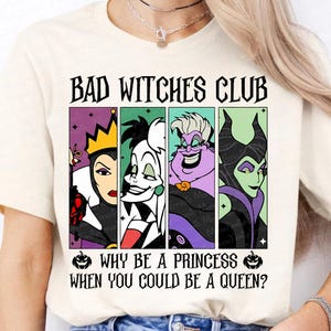 May include: Cream-colored t-shirt featuring the text "BAD WITCHES CLUB" above a graphic of four cartoon villains. Below the graphic, the text reads "WHY BE A PRINCESS WHEN YOU COULD BE A QUEEN?" The shirt has short sleeves.