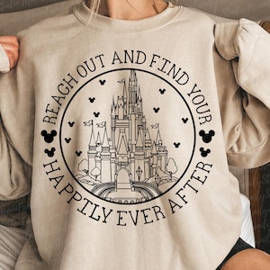 以下が含まれることがあります： ベージュのトレーナーに、黒い城のグラフィックプリントと「Reach out and find your happily ever after」というテキストがプリントされています。