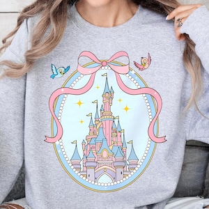 May include: A light gray sweatshirt featuring a fairytale castle design. The castle is pink and blue, surrounded by a pink bow and a pearl-like border. Two cartoon birds are on either side of the bow.