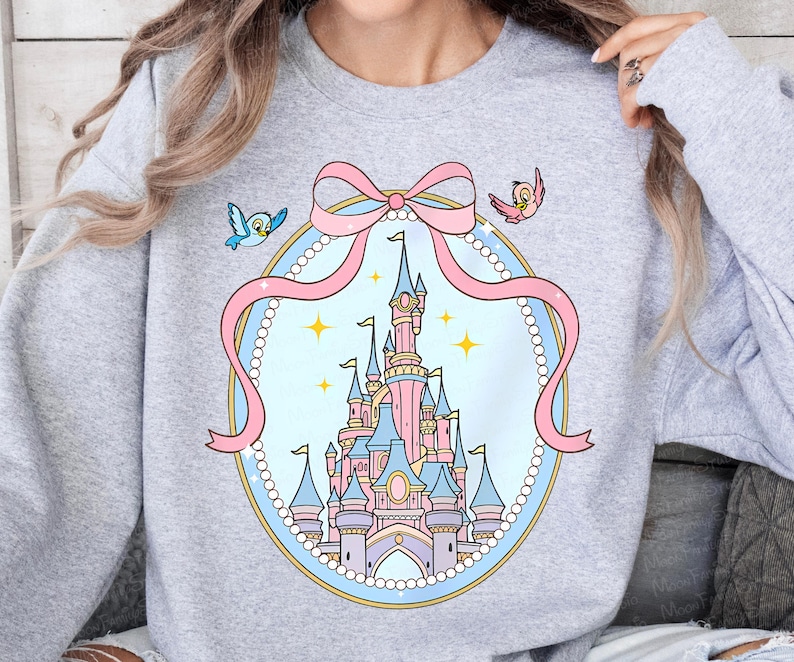 May include: A light gray sweatshirt featuring a fairytale castle design. The castle is pink and blue, surrounded by a pink bow and a pearl-like border. Two cartoon birds are on either side of the bow.