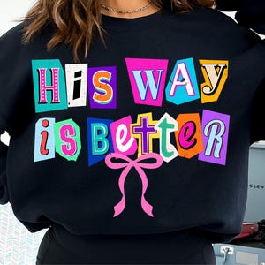 May include: Black sweatshirt with the text "His Way is Better" in colorful, cut-out style letters. A pink bow graphic is below the text. The sweatshirt is a casual, comfortable style.