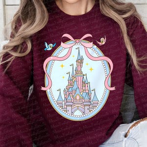 May include: Burgundy sweatshirt featuring a whimsical illustration of a fairytale castle within a pink ribbon frame. Two small birds are perched on either side of the ribbon. The design is set against a burgundy background.