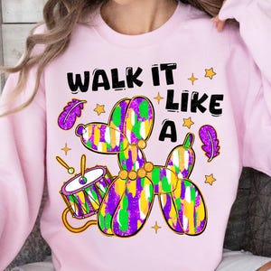 May include: A light pink sweatshirt with a colorful balloon dog design. The design includes a drum, stars, and the text "WALK IT LIKE A". The balloon dog is purple, green, and yellow.