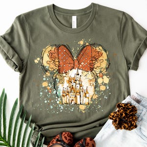 May include: Olive green t-shirt featuring a whimsical design. The design includes a polka-dot bow, Mickey Mouse ears, and a castle illustration. The graphic is surrounded by colorful paint splatters, creating a playful and artistic look.