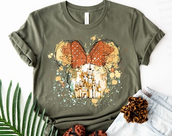 May include: An olive green t-shirt with a playful design. The design features a polka-dot bow, Mickey Mouse ears, and a castle illustration. The graphic is surrounded by colourful paint splatters, creating an artistic look.