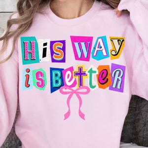 May include: A light pink sweatshirt with the phrase "His Way is Better" in a colorful, cut-out style design. The text is in various colors, including pink, blue, orange, and yellow, with a pink bow at the bottom.