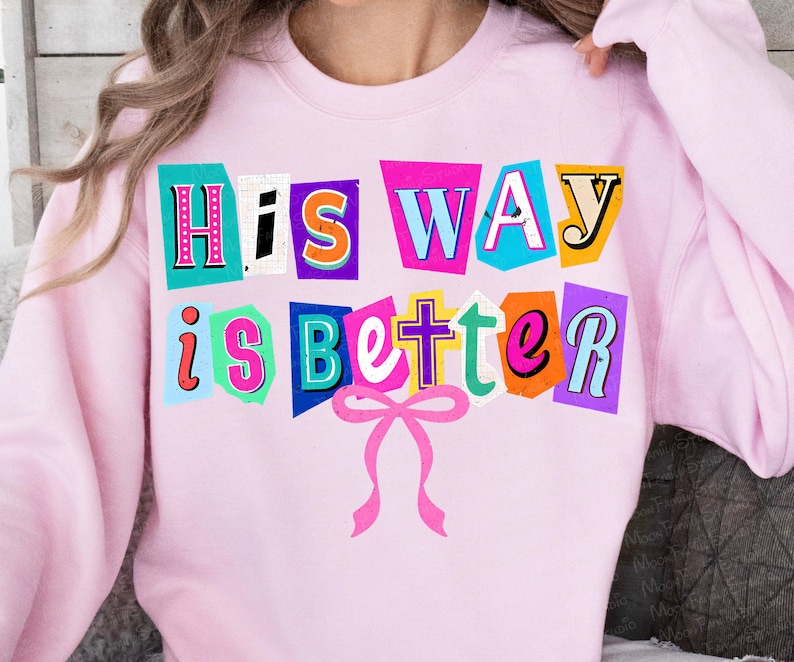 May include: A light pink sweatshirt with the phrase "His Way is Better" in a colorful, cut-out style design. The text is in various colors, including pink, blue, orange, and yellow, with a pink bow at the bottom.