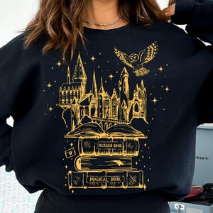 Könnte beinhalten: Schwarzes Sweatshirt mit einer goldenen Illustration eines Schlosses, einer Eule und Büchern. Die Bücher sind mit "Wizard Book" und "Magical Book" beschriftet. Das Design enthält Sterne und einen Brief. Eine goldene Halskette ist sichtbar.