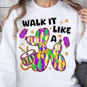 May include: White sweatshirt with the text "WALK IT LIKE A" above a colorful balloon animal dog, drum, stars, and feathers. The balloon animal is purple, green, and yellow. The drum is purple, gold, and white.