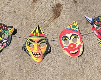 Puede incluir: Una cadena de siete máscaras de Halloween con varios personajes, incluyendo una bruja, un diablo, un payaso, un gato y un zombi. Las máscaras son coloridas y tienen un aspecto vintage.