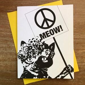 May include: A black and white greeting card featuring a cat with a floral headpiece holding a sign with a peace symbol and the word "MEOW!". The card is paired with a yellow envelope, perfect for sending a message of peace and feline fun.