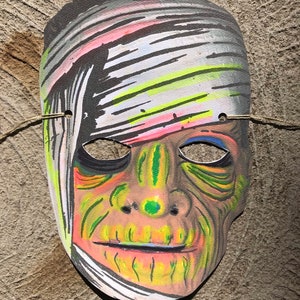 Op de afbeelding: Een papieren Halloween-masker van een mummie met een groen en geel gezicht en witte banden. Het masker heeft een gat voor de ogen en een mond.