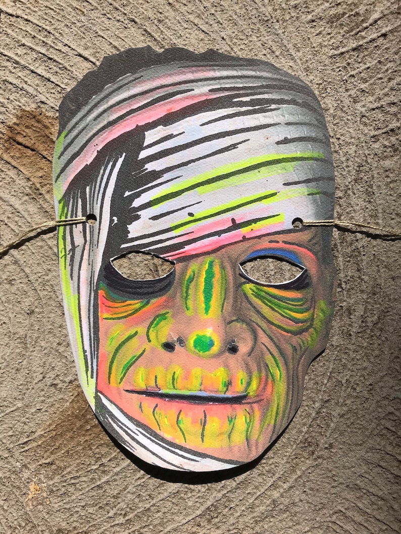 Op de afbeelding: Een papieren Halloween-masker van een mummie met een groen en geel gezicht en witte banden. Het masker heeft een gat voor de ogen en een mond.