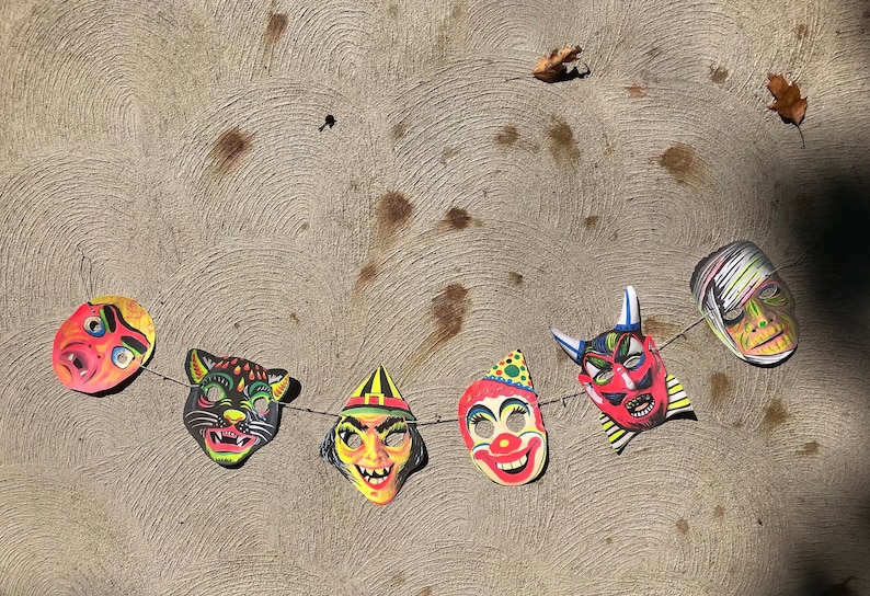Op de afbeelding: Een slinger met zeven Halloween-maskers met verschillende personages, waaronder een heks, een duivel, een clown, een kat en een zombie. De maskers zijn kleurrijk en hebben een vintage uitstraling.