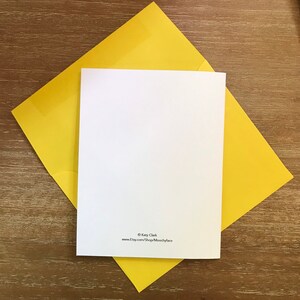 May include: A blank white greeting card with rounded corners rests on a bright yellow envelope. The card and envelope are on a wooden surface. The card has text at the bottom that reads "&copy; Katy Clark" and "www.etsy.com/Shop/Moochyface".