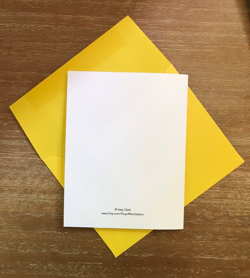 May include: A blank white greeting card with rounded corners rests on a bright yellow envelope. The card and envelope are on a wooden surface. The card has text at the bottom that reads "&copy; Katy Clark" and "www.etsy.com/Shop/Moochyface".