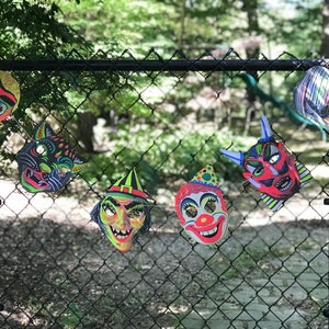 Op de afbeelding: Een verzameling van zes vintage Halloween-maskers die aan een zwart gaashek hangen. De maskers zijn gemaakt van karton en tonen verschillende personages, waaronder een heks, een clown, een duivel, een kat en twee andere griezelige gezichten. De maskers zijn felgekleurd en hebben een retro-look.