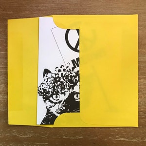May include: A yellow envelope partially open, revealing a black and white art print. The print features a stylized cat face with floral elements and the word "M" visible. The background is a wooden surface.