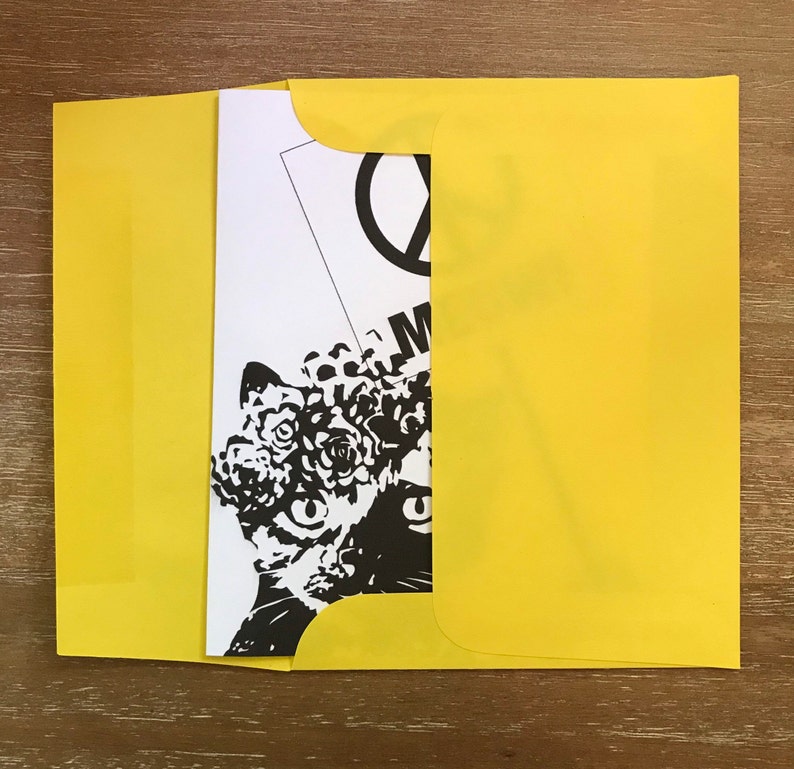 May include: A yellow envelope partially open, revealing a black and white art print. The print features a stylized cat face with floral elements and the word "M" visible. The background is a wooden surface.
