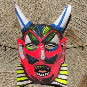 Op de afbeelding: Een rood duivels masker met blauwe en witte hoorns, zwarte ogen en een geel-zwarte gestreepte kraag. Het masker heeft een dreigende uitdrukking met een wijd open mond en scherpe tanden.