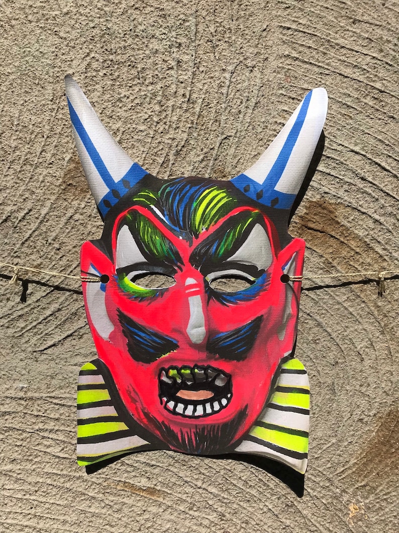 Op de afbeelding: Een rood duivels masker met blauwe en witte hoorns, zwarte ogen en een geel-zwarte gestreepte kraag. Het masker heeft een dreigende uitdrukking met een wijd open mond en scherpe tanden.