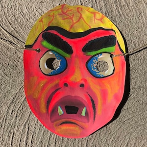 Op de afbeelding: Een rood en geel papieren masker met een eng gezicht. Het masker heeft een brede mond met scherpe tanden, gefronste wenkbrauwen en grote blauwe ogen met zwarte pupillen. Het masker heeft touwtjes om vast te binden.