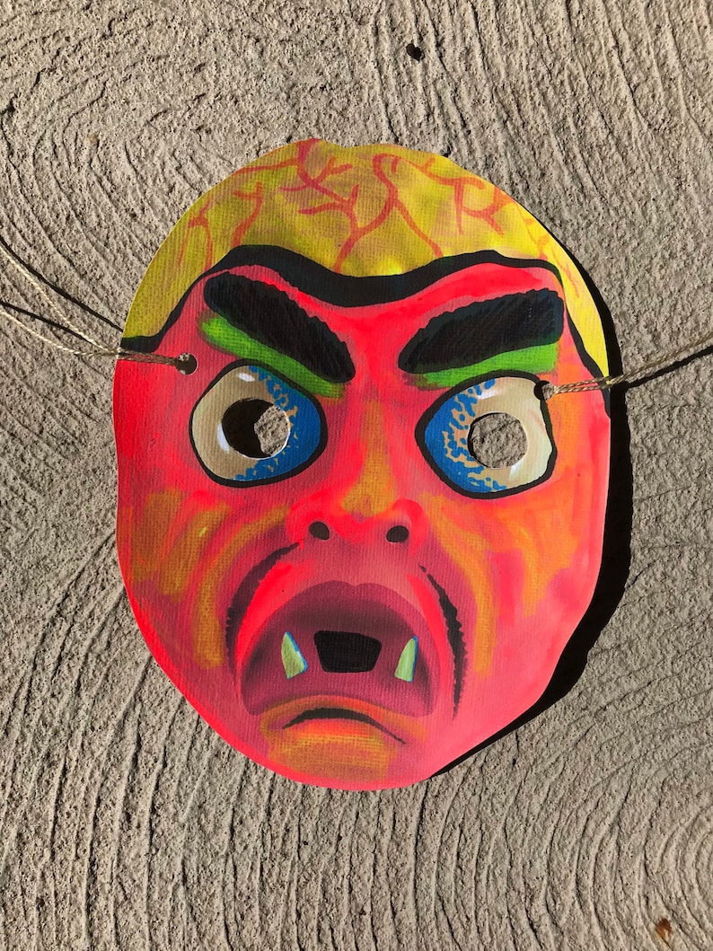 Op de afbeelding: Een rood en geel papieren masker met een eng gezicht. Het masker heeft een brede mond met scherpe tanden, gefronste wenkbrauwen en grote blauwe ogen met zwarte pupillen. Het masker heeft touwtjes om vast te binden.