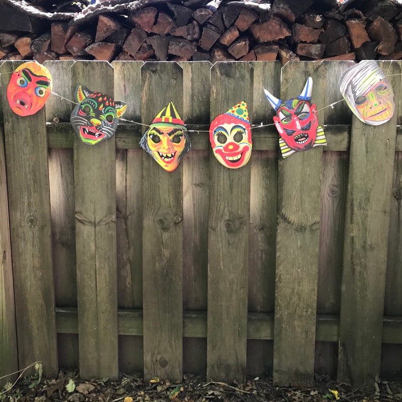 Op de afbeelding: Een reeks van zes Halloween-maskers die aan een houten hek hangen. De maskers zijn gemaakt van papier en tonen verschillende personages, waaronder een clown, een duivel, een kat en een monster.