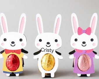 May include: Three white bunny-shaped cardboard boxes with cut-out windows to hold chocolate eggs. The bunny on the left is wearing a yellow shirt with a red bow tie. The bunny in the middle has the name "Cristy" written on its chest and is wearing a gold foil-wrapped chocolate egg. The bunny on the right is wearing a purple dress with a pink bow.