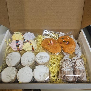 May include: A box filled with assorted baked goods. The box contains meringue cookies, orange-colored cookies with sprinkles, and powdered sugar cookies. Two small bags of chocolate candies are also included, all nestled on yellow paper shreds.