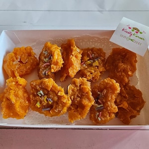 May include: A white cardboard box with a white paper doily inside. The box contains eight orange, crispy, fried treats with colorful sprinkles on top. The box has a small pink and green sticker with the text "Kristy Kramer" on it.