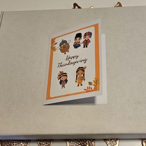 May include: A white box with a card inside. The card has an orange background with a happy Thanksgiving message and a cartoon illustration of people in traditional clothing.