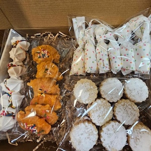 May include: A white gift box filled with various baked goods. The box contains three types of cookies: meringue cookies with colorful sprinkles, orange cookies with colorful sprinkles, and shortbread cookies dusted with powdered sugar. The cookies are packaged in clear plastic bags.