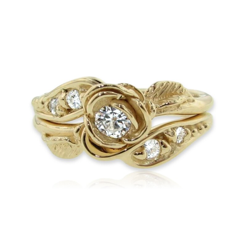 Front view of yellow gold wedding set with a rose bloom center. The rose is made up of five petals that curve around a diamond. A leaf curves around the band of the engagement ring, and the wedding band has small diamonds set low into the design