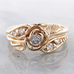 Front view of yellow gold wedding set with a rose bloom center. The rose is made up of five petals that curve around a diamond. A leaf curves around the band of the engagement ring, and the wedding band has small diamonds set low into the design