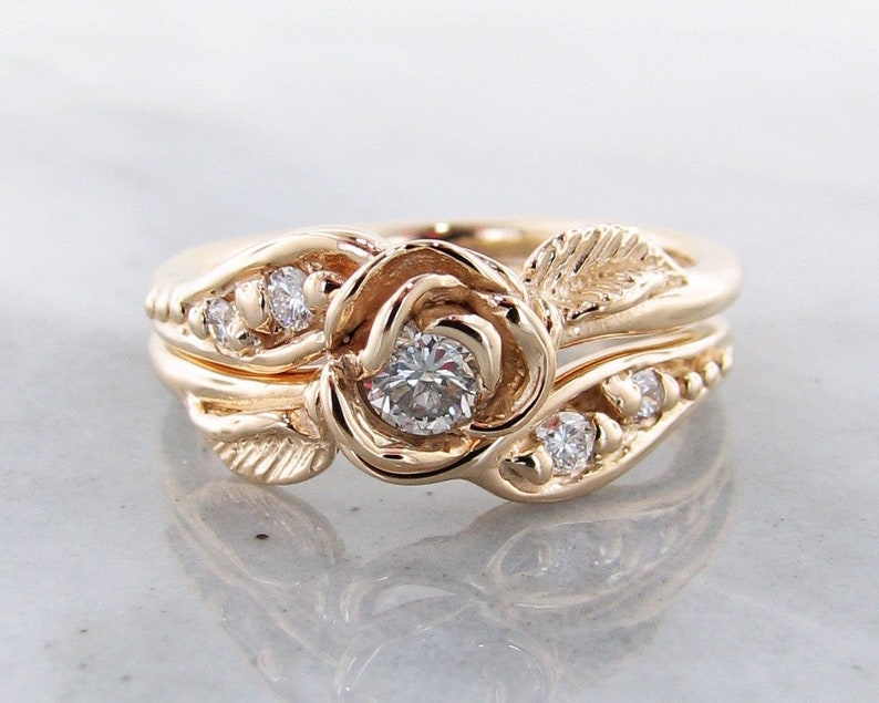 Front view of yellow gold wedding set with a rose bloom center. The rose is made up of five petals that curve around a diamond. A leaf curves around the band of the engagement ring, and the wedding band has small diamonds set low into the design