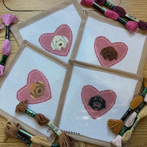 May include: Four square needlepoint canvases with a pink heart design, each featuring a different dog breed. The canvases are bordered with a tan fabric and surrounded by spools of embroidery floss in various shades of pink and brown.