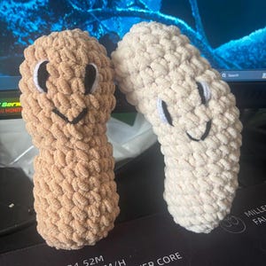 May include: Two crocheted peanut-shaped plush toys. One is brown and the other is white. Both have embroidered eyes and a smiling mouth.