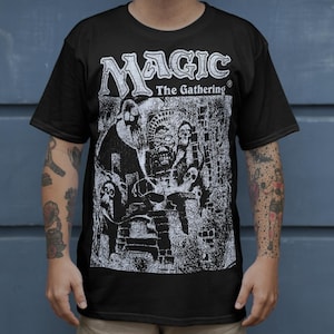 Puede incluir: Camiseta negra con el texto "MAGIC The Gathering" en una fuente estilizada. La camiseta presenta un gráfico detallado en blanco y negro de una escena de fantasía con figuras y elementos arquitectónicos. La persona lleva pantalones cortos color caqui.