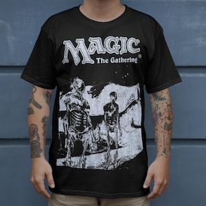 May include: Black t-shirt with the "Magic: The Gathering" logo and a monochrome graphic of skeletal figures. The shirt has short sleeves and a crew neck. The person is wearing beige shorts.