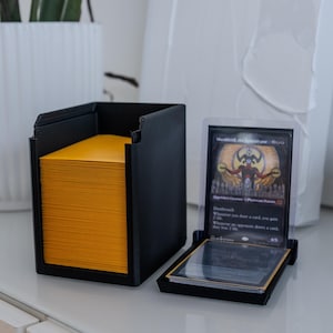 May include: A black card storage box with a yellow deck of cards inside. A black card holder with a clear plastic sleeve containing a black and red "Dreadnought,  Dreadknight" Magic: The Gathering card is next to the box.